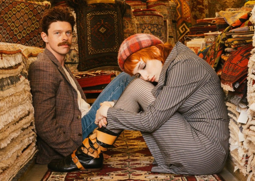 Two people sat on the floor surrounded by piles of rugsx